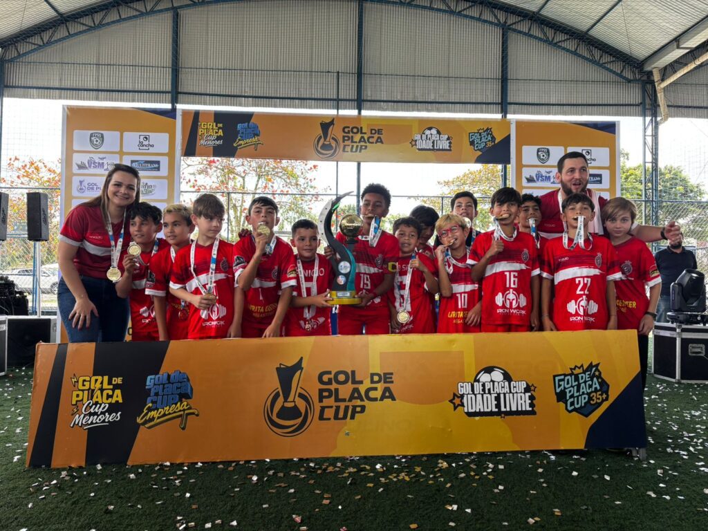 "Foto da equipe de futebol Sub-10 da Escola Rubra de Pontal do Paraná posando com o troféu de campeão. Os jovens atletas e a comissão técnica sorriem para a câmera, exibindo suas medalhas douradas em frente ao banner do evento Gol de Placa Cup."