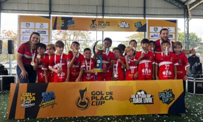 "Foto da equipe de futebol Sub-10 da Escola Rubra de Pontal do Paraná posando com o troféu de campeão. Os jovens atletas e a comissão técnica sorriem para a câmera, exibindo suas medalhas douradas em frente ao banner do evento Gol de Placa Cup."
