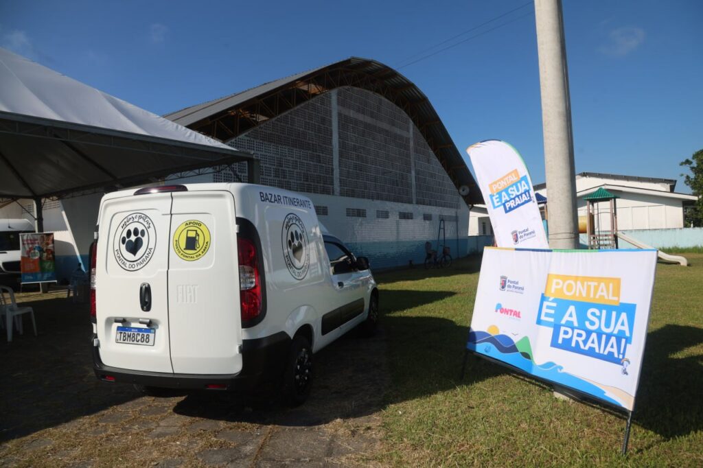 Veículo utilitário branco da Rede de Proteção Animal e placa informativa da campanha municipal de Pontal do Paraná estacionados em frente ao Ginásio da Escola Amatuzzi.